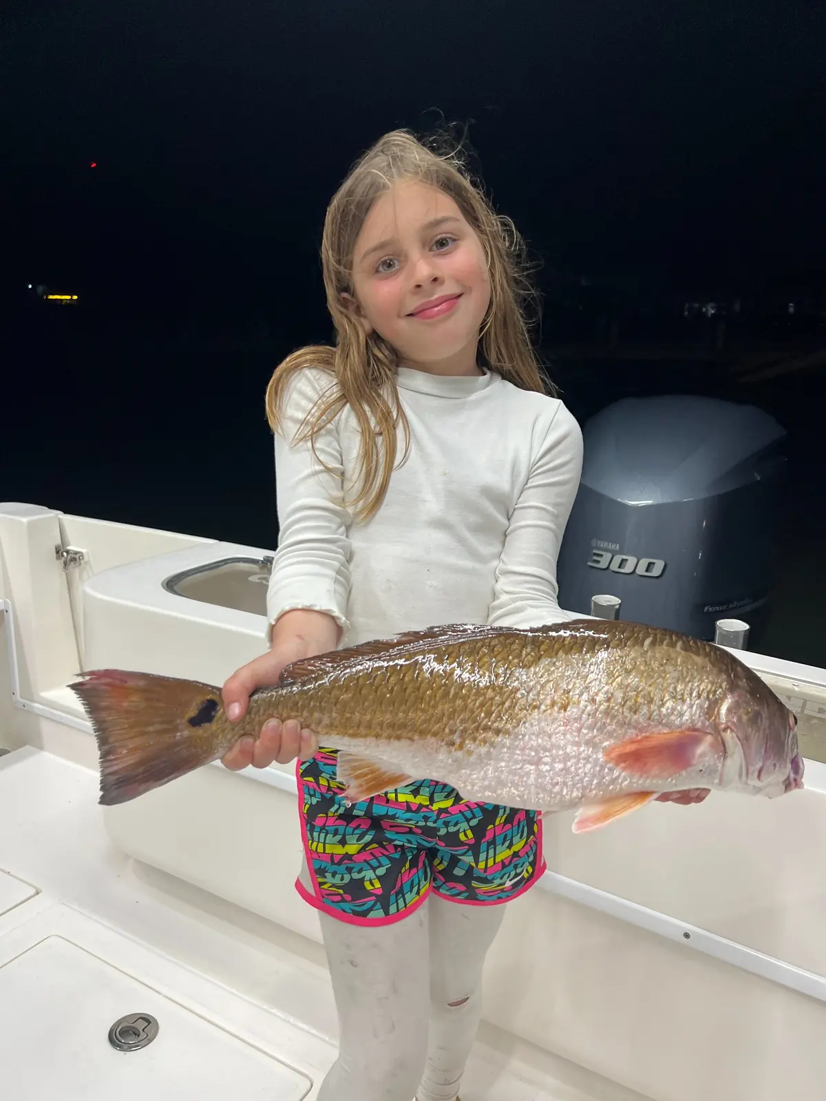 A child holds a large redfish aboard a boat at night.